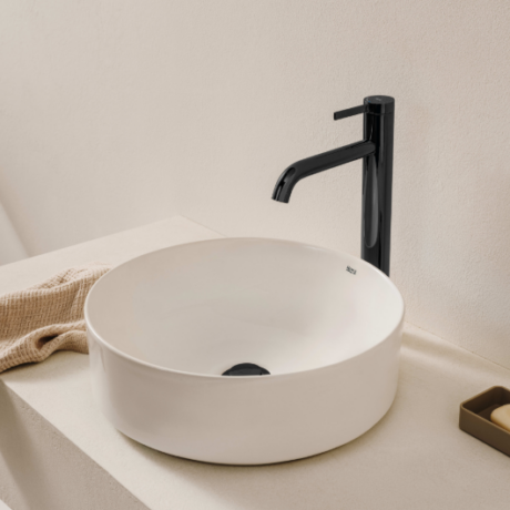 White round vessel sink with a tall black faucet on a beige countertop, with a towel and soap tray nearby.