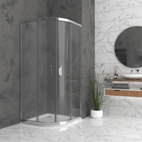 Curved glass shower enclosure with chrome frame in a marble-tiled bathroom, next to a potted plant and a floating vanity with a round mirror.