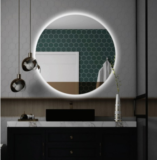 Round backlit mirror above a marble-topped bathroom vanity with a black sink, dark cabinetry, hexagonal wall tiles, bronze pendant lights, and a vase on the counter.
