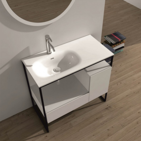 Modern white bathroom vanity with sink and open shelf, black metal frame, round mirror above, on wooden floor with a stack of books nearby.