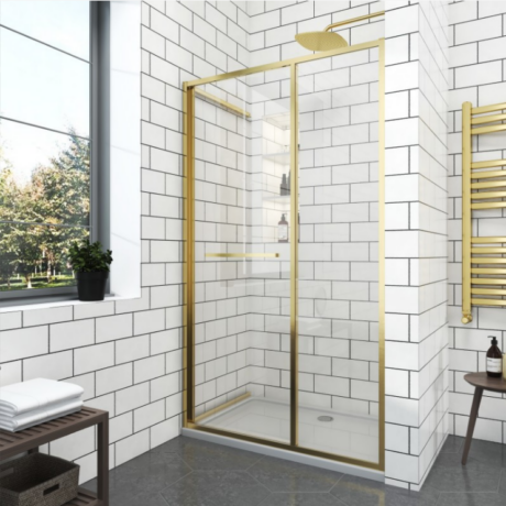 Gold-framed glass shower enclosure in a modern white-tiled bathroom with a rainfall showerhead and a small shelf inside.